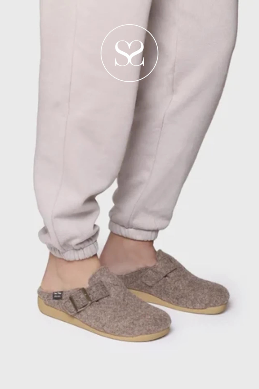 A person wearing light beige jogger pants and taupe-brown felt clogs with buckle straps, shown from the knees down against a plain light gray studio background. A circular 'SC' logo watermark is visible near the upper leg.