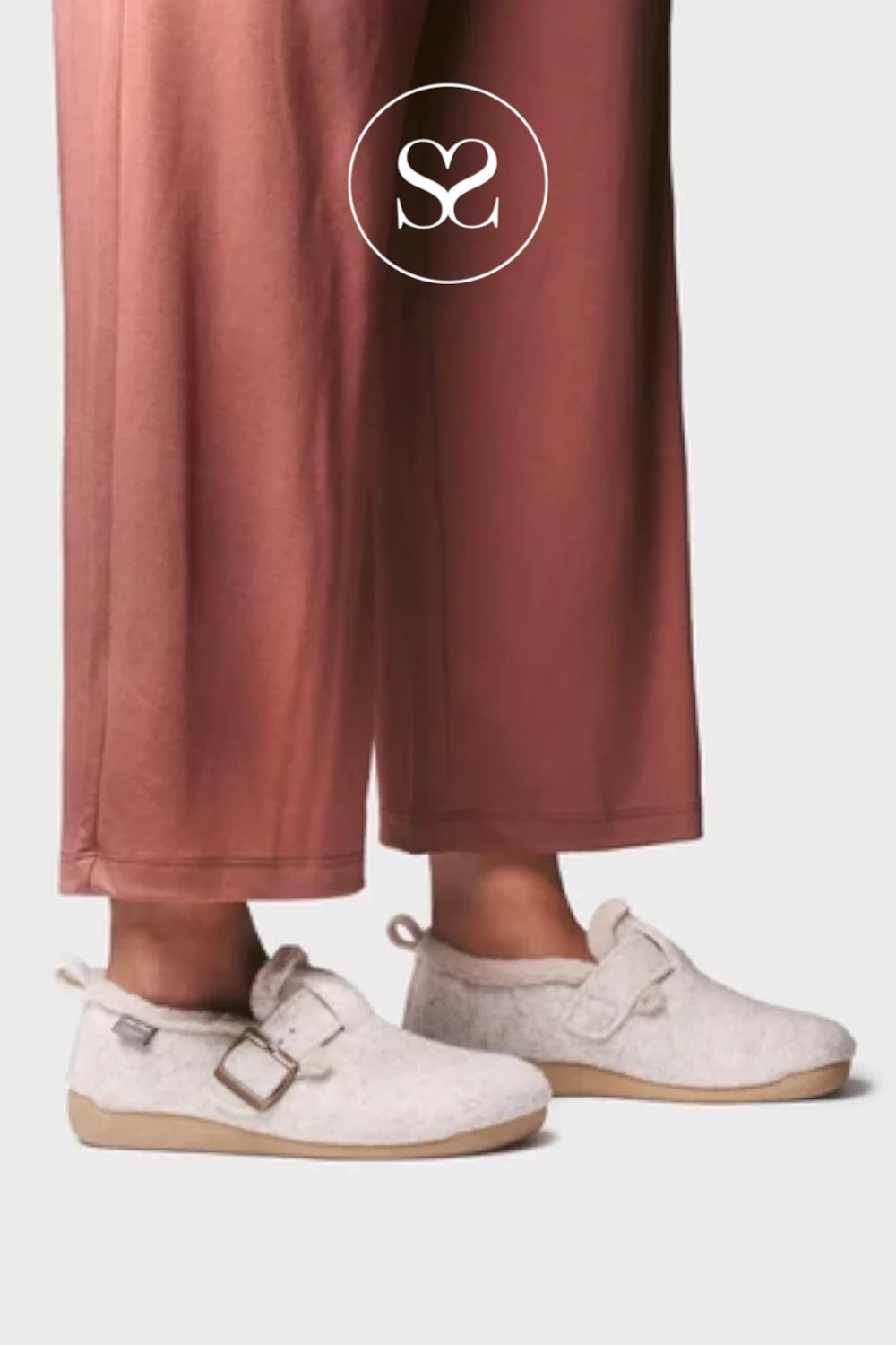A close-up of a person's feet wearing light beige slip-on clog-style shoes with buckle straps, paired with dusty rose wide-leg pants against a light gray background. A white circular logo with an 'S' and '2' is overlaid near the top.