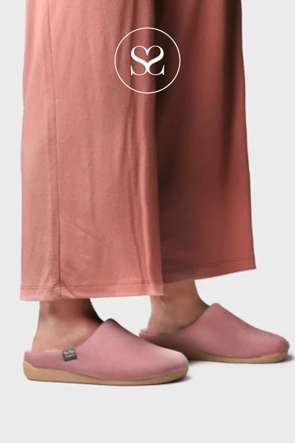 A person wearing dusty pink slip-on clogs and matching wide-leg trousers, shown from the calves down against a light gray studio background. A white circular logo with an 'S' mark is overlaid near the top.