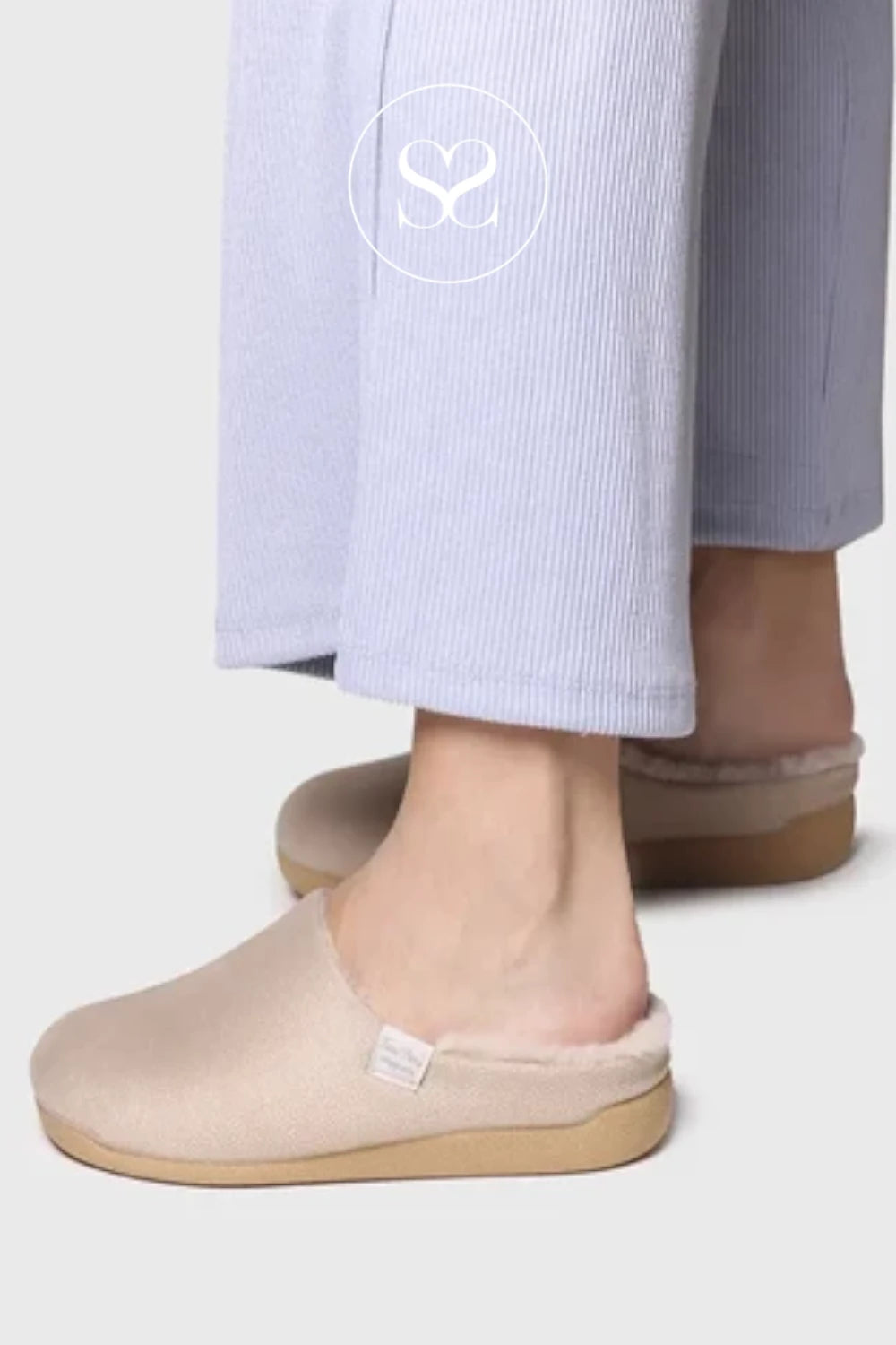 A close-up of a person wearing light beige slip-on slippers with a soft fuzzy lining and light brown soles, paired with pale lavender ribbed trousers. A white circular logo with an 'S' is visible on the pant leg.