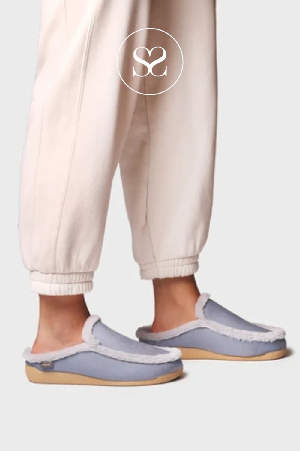 A person wearing light beige jogger pants and light blue slip-on slippers with white faux-fur trim, shown from the calves down against a plain light gray background. A white circular 'S2' logo is overlaid near the upper center.
