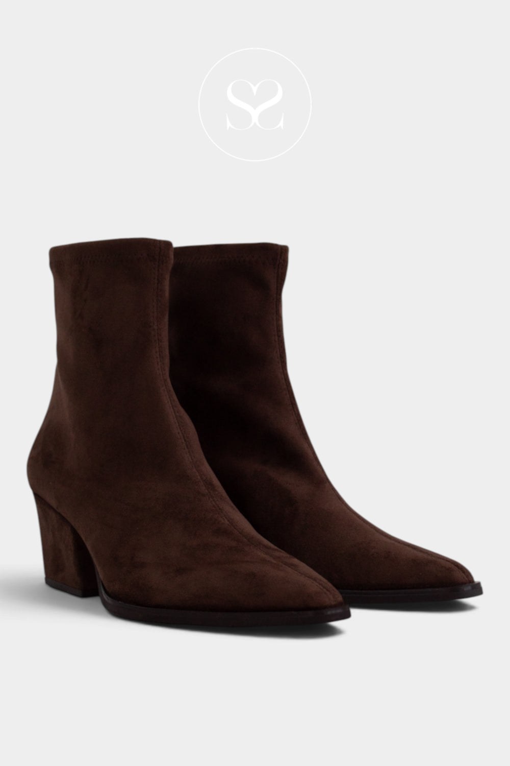 A pair of dark brown suede ankle boots with pointed toes and low block heels, displayed against a light gray background with a faint circular logo at the top.