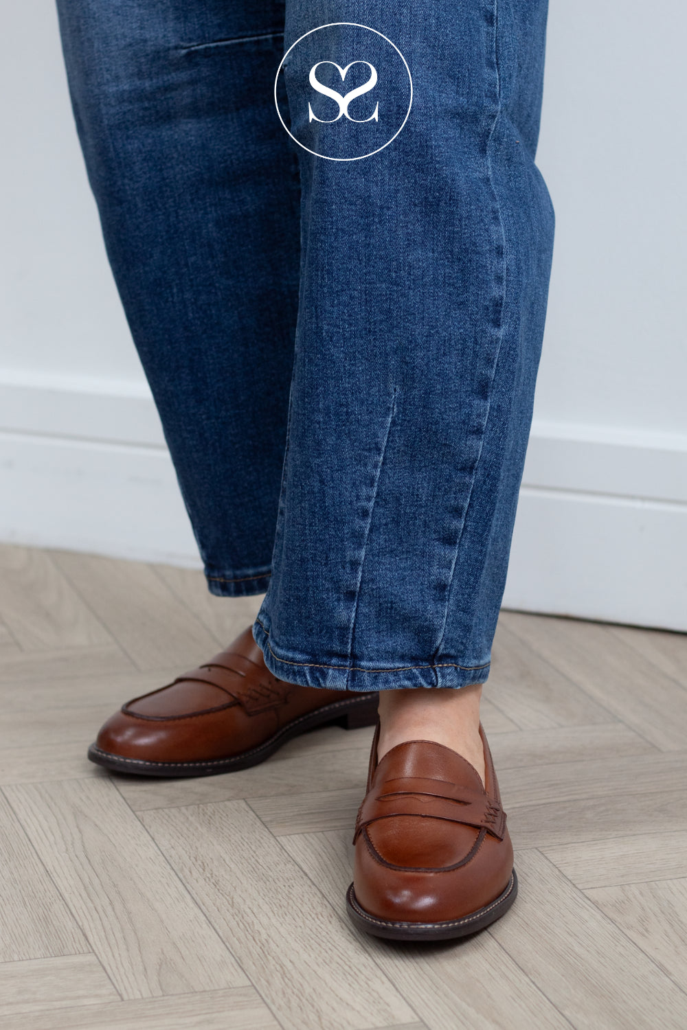 A woman wearing blue jeans and classic brown leather penny loafers from Tamaris standing on a light wood herringbone floor against a white wall.