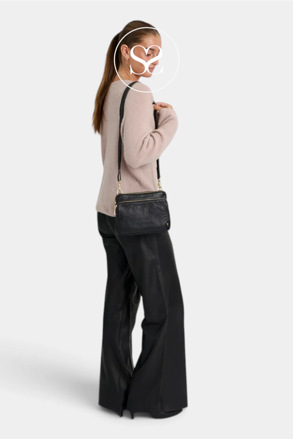 A woman shown from the back wearing a light beige long-sleeve top, black wide-leg pants, and carrying a black crossbody bag with gold-tone hardware against a light gray studio background.