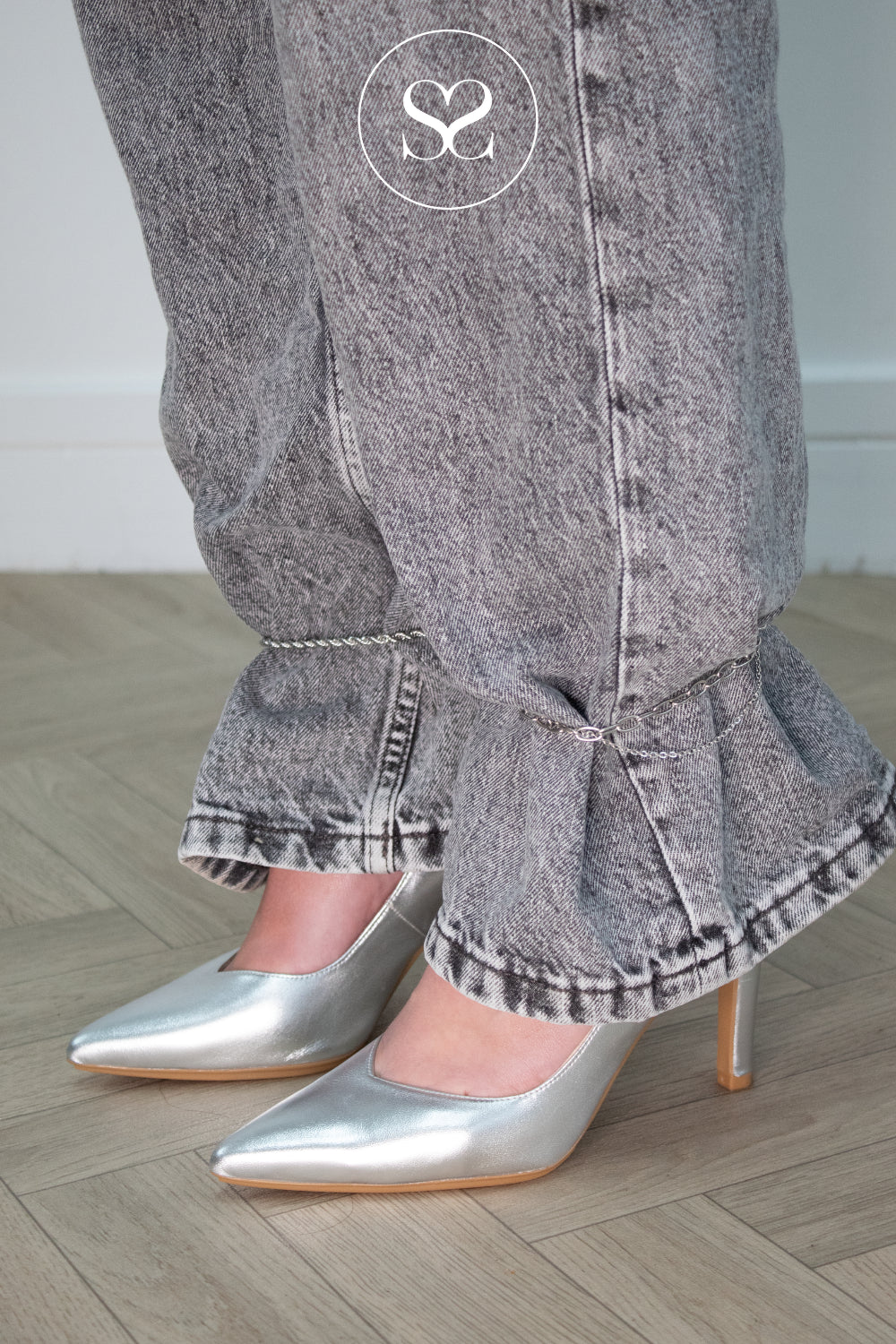 Person wearing silver high-heeled stilettos from lodi with gray flared jeans on a wooden floor.