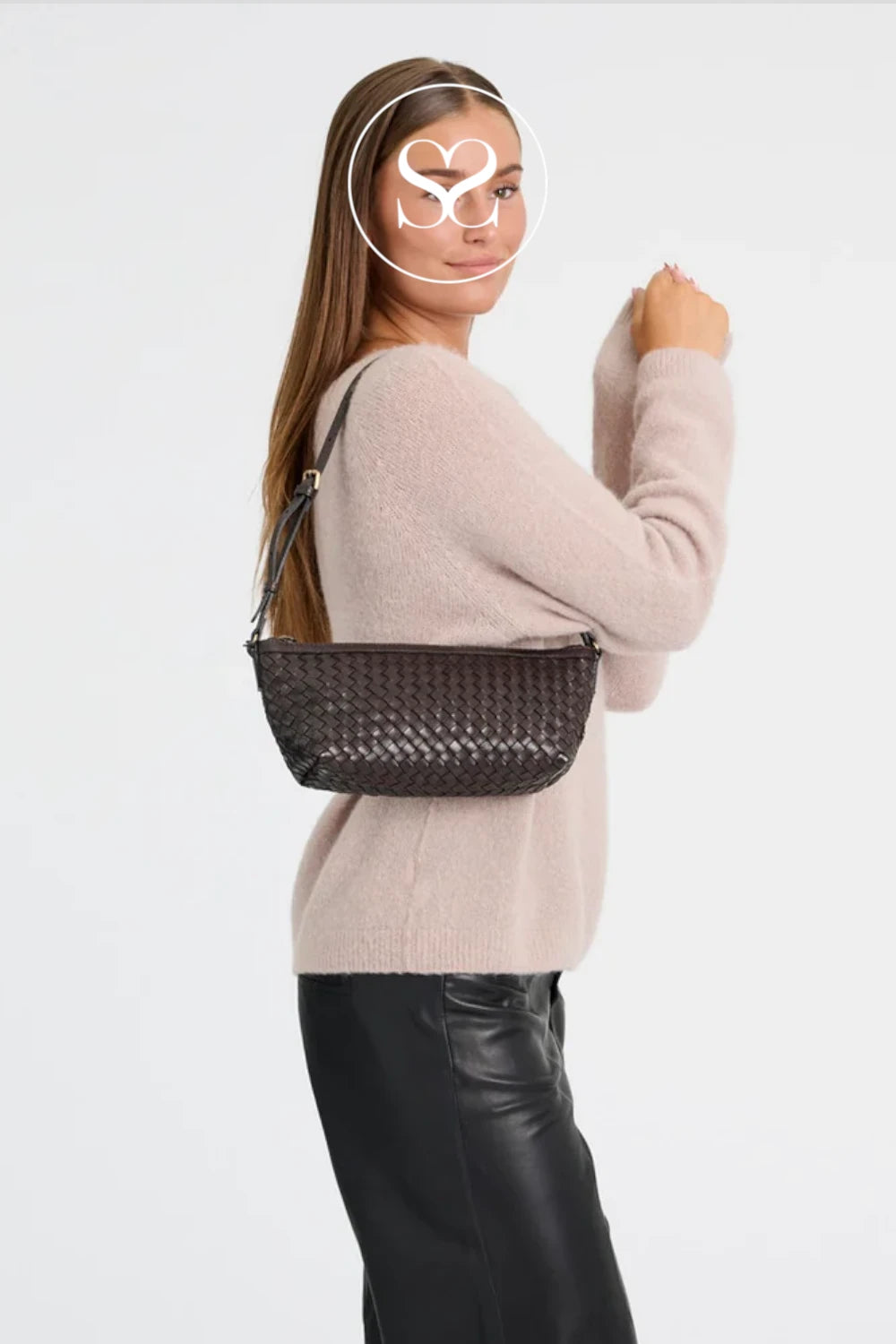 A woman wearing a light pink knit sweater and black leather pants carries a dark brown woven basket-style shoulder bag against a plain light gray studio background.