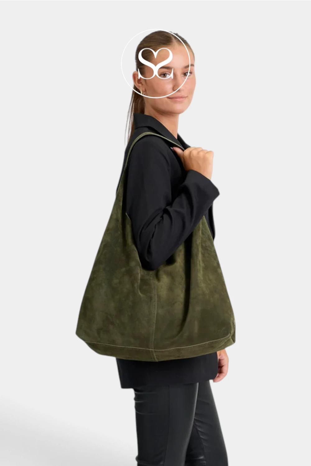 A woman wearing a black long-sleeve top and black pants carries an olive green suede-like shoulder bag against a light gray studio background. A white circular logo overlay is visible near the top of the image.