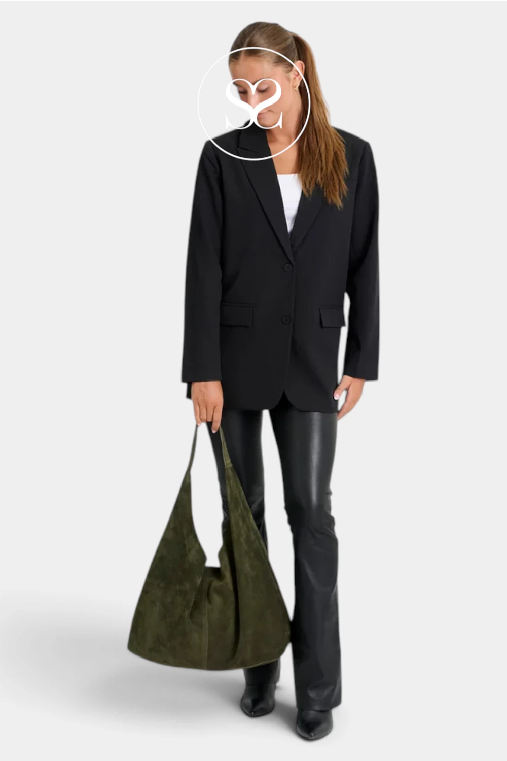 A model wearing a black blazer, black leather pants, and black boots, holding an olive green suede slouchy shoulder bag against a light gray background.