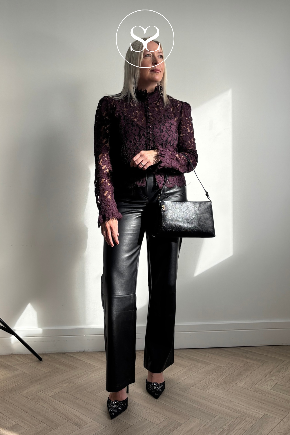 A woman stands in a room wearing a dark burgundy lace long-sleeve top, black leather pants, Tamaris black pointed party heels, and carrying a black textured shoulder bag from Katie Loxton. A white circular logo overlay is centered on her face.