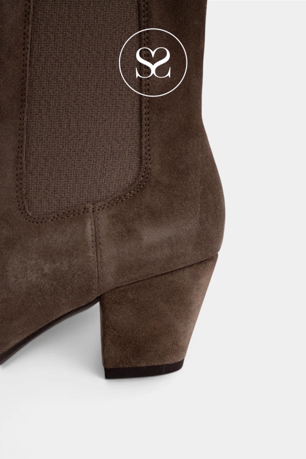 Close-up of the heel and side panel of a Unisa Taupe brown suede boot with a block heel and elastic side gore, shown on a light gray background with a white circular Sheneil Shoes logo overlay.