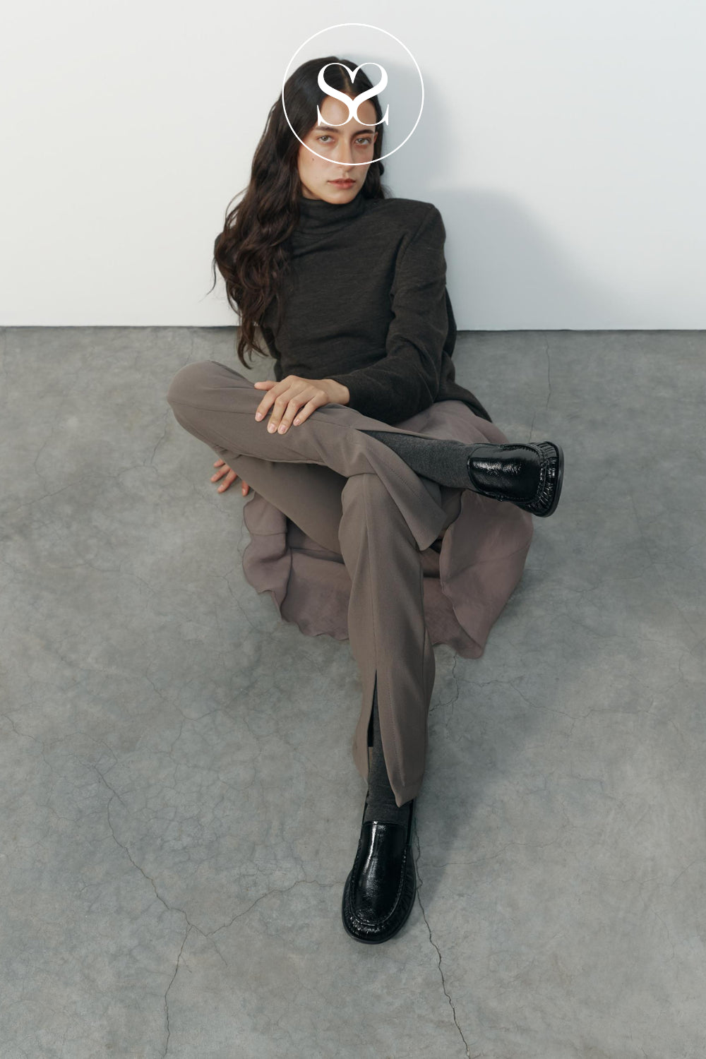 A model sits cross-legged on a gray concrete floor against a white wall, wearing a dark brown turtleneck, taupe wide-leg trousers, and blackhigh shine rouched loafers from Vagabond shoemakers. A white circular sheneil shoes logo overlay is positioned over her face.