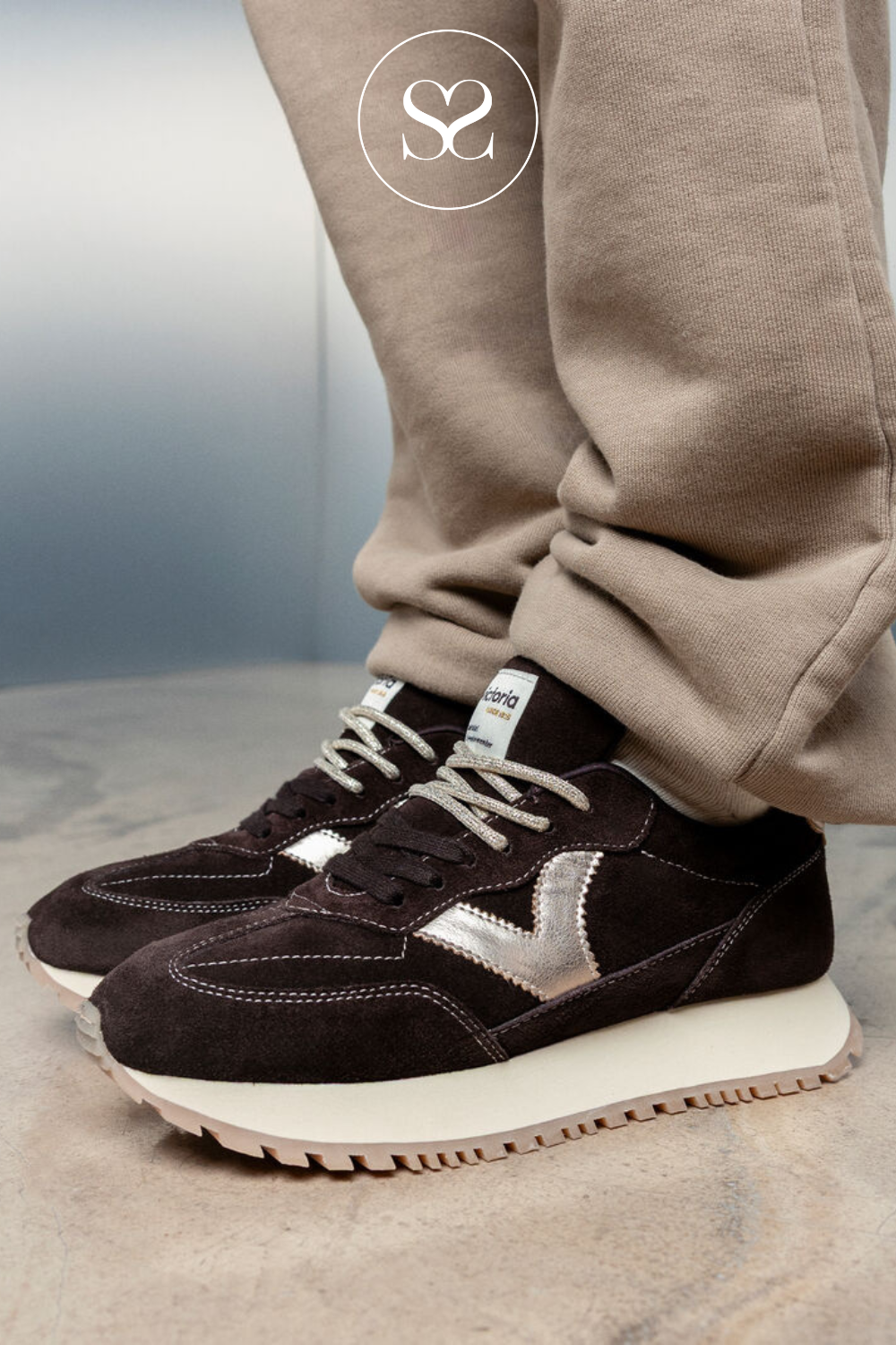 A close-up of a person's legs wearing dark brown suede sneakers with metallic silver side stripes and beige sweatpants, standing on a textured stone floor.