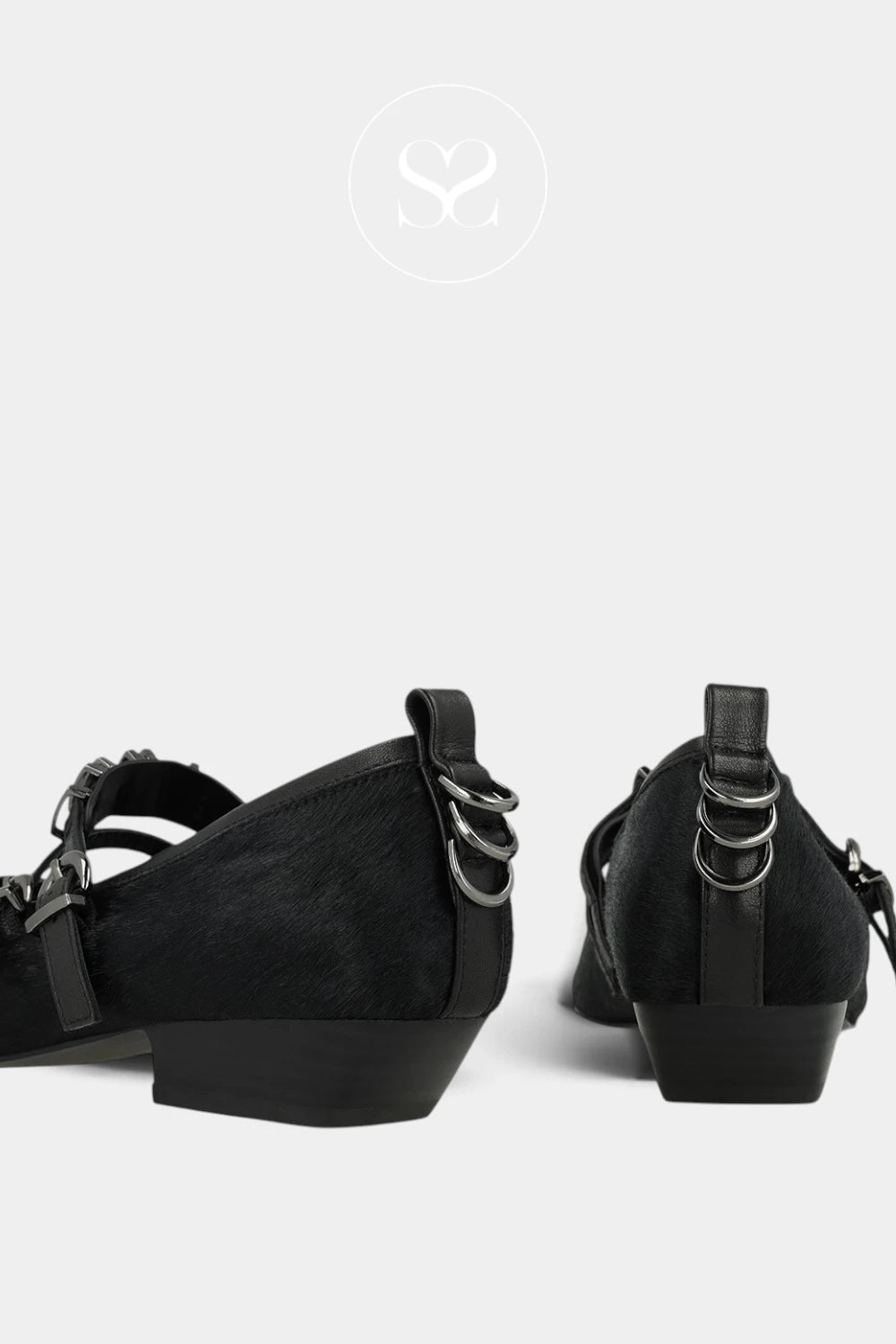 A pair of black wedge shoes shown from the back, featuring textured black uppers, black straps with silver-tone metal rings, and chunky black wedge heels, set against a light gray background.