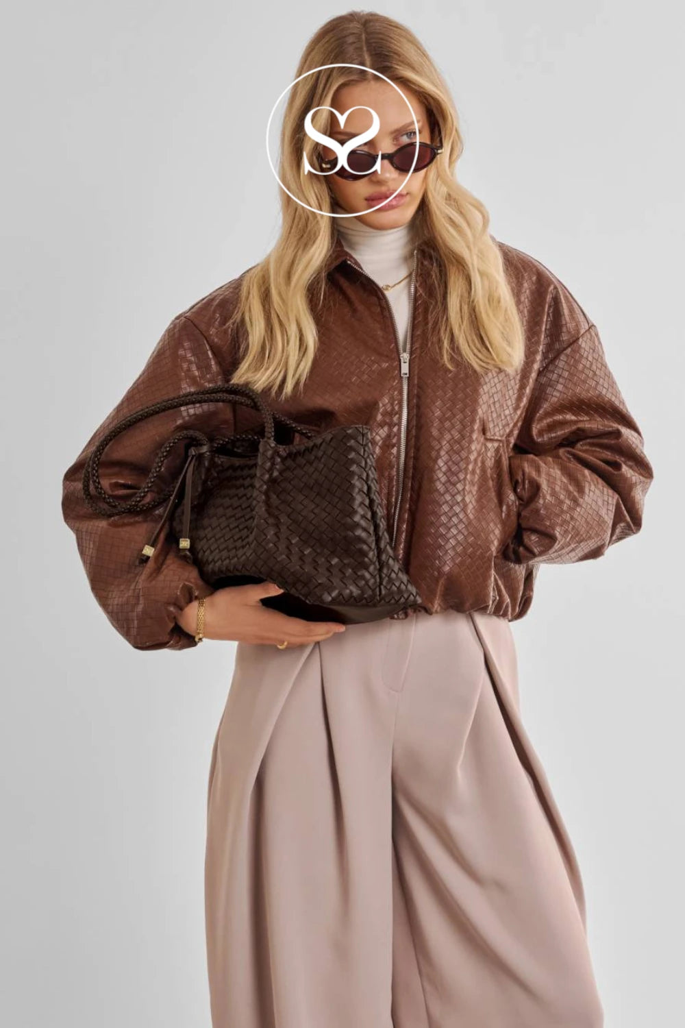 A woman wearing a brown textured bomber jacket, light pink wide-leg trousers, and sunglasses, holding a dark brown woven handbag against a light gray studio background.