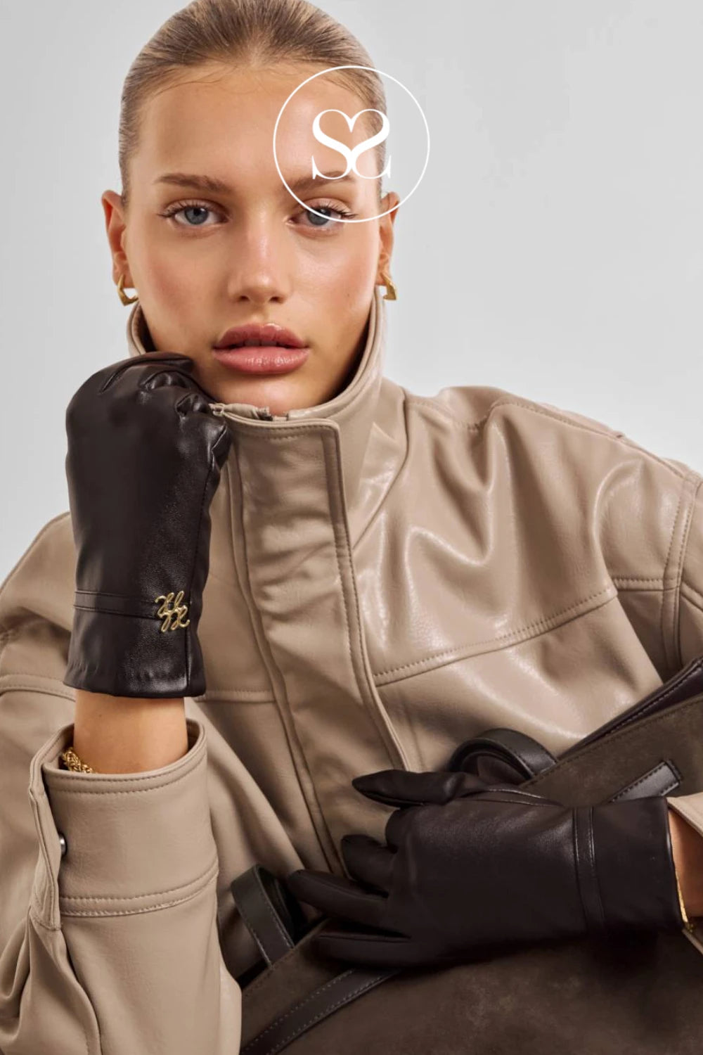 A model wearing a beige faux-leather jacket and black leather gloves with gold hardware, posed against a light gray studio background.