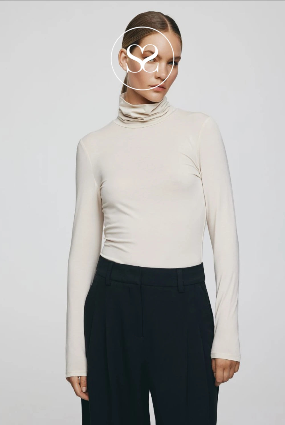 A model wearing a fitted, long-sleeve cream-colored cowl neck top paired with dark trousers, set against a plain light gray studio background.