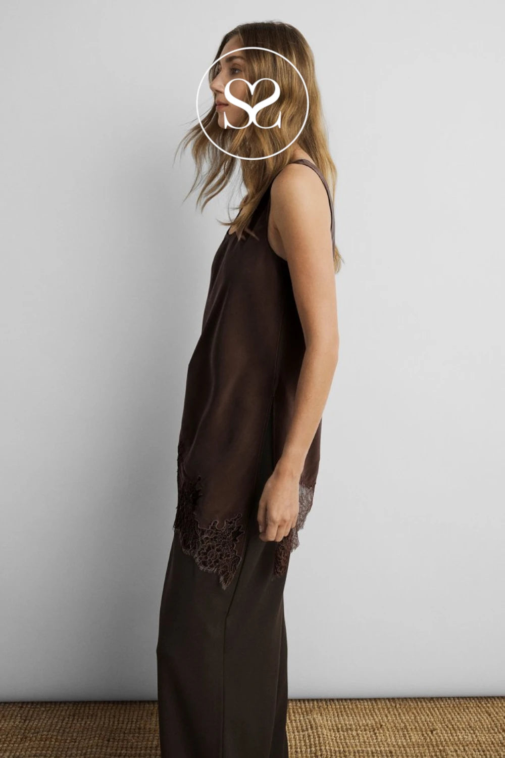 A woman wearing a dark brown sleeveless slip dress with lace trim at the hem, standing against a light gray wall.