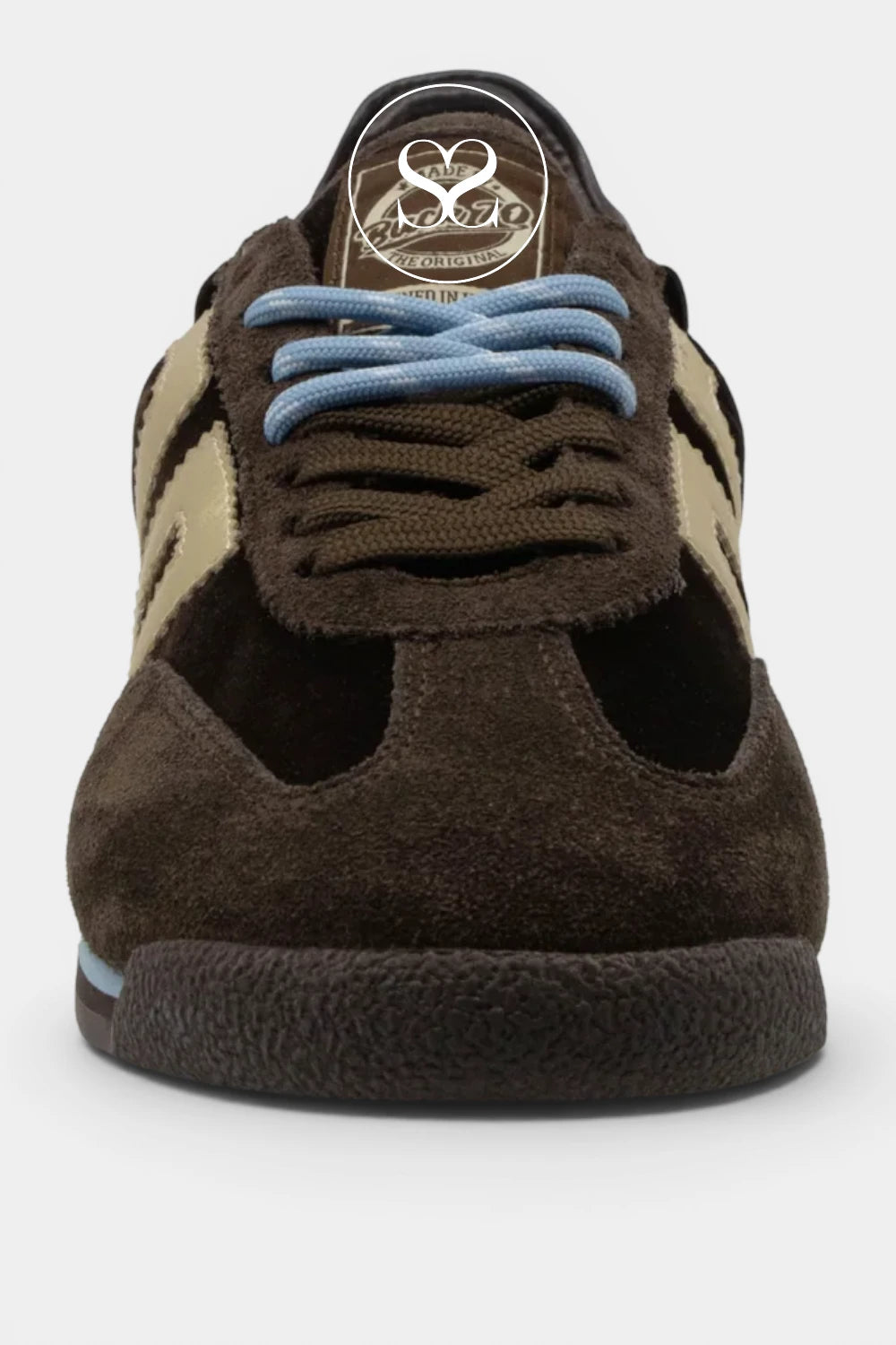 A close-up shot of a brown suede and leather sneaker, showing the side profile with light blue laces, beige side stripes, and a circular metallic logo patch on the tongue.