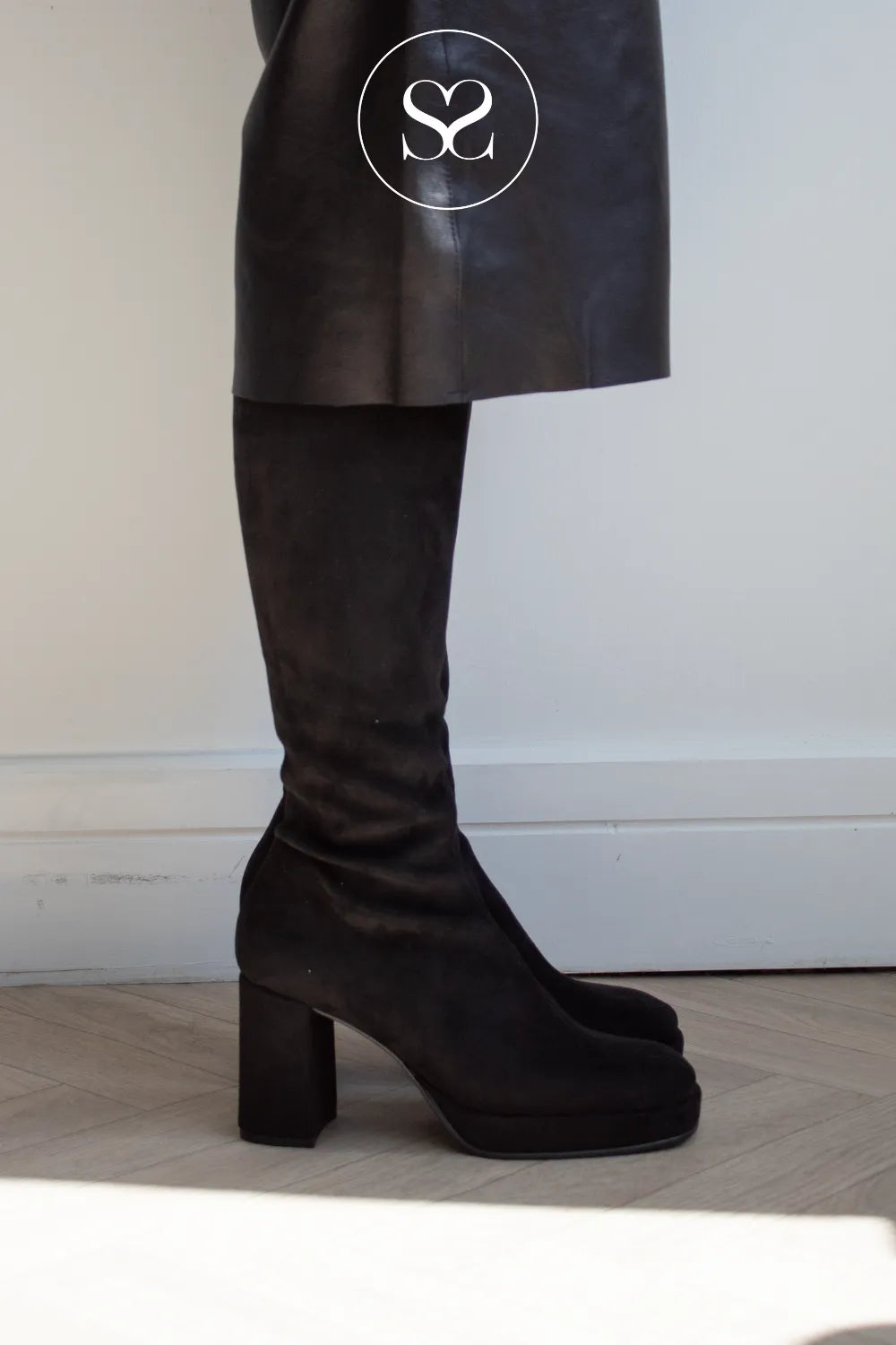 A person wearing a black leather skirt and a black suede knee high boot with a chunky block heel from Miss Elastic, standing on a light wood floor against a white wall.