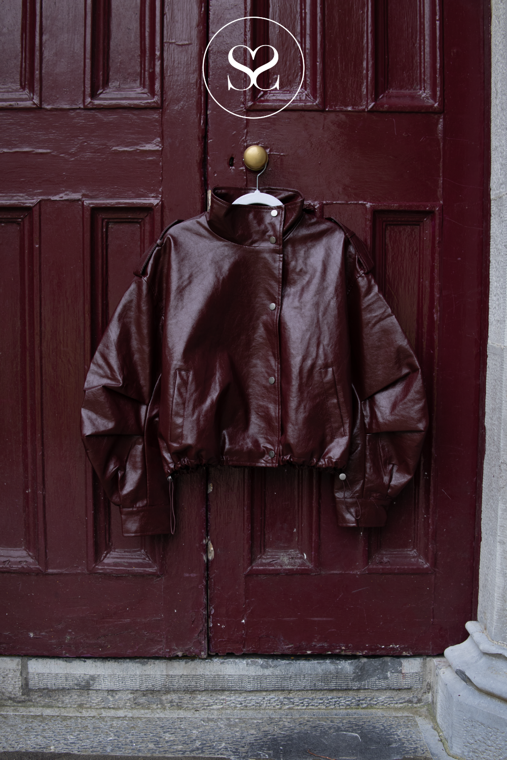 A glossy dark burgundy cropped jacket hanging on a hanger against a dark red wooden door, with a circular 'S2' logo overlay at the top.