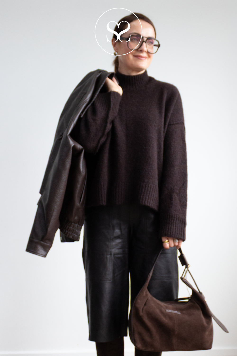 Editorial Image of A woman wearing a dark brown knit turtleneck sweater, black leather pants, and glasses, carrying a black leather jacket and holding a Depeche Chocolate brown suede hobo bag against a light grey studio background in Ireland