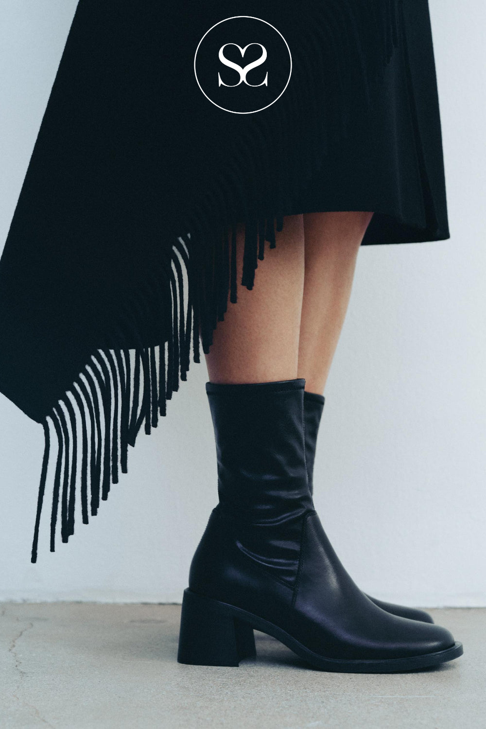 A person wearing black leather mid-calf sock boots with a block heel and round toe from Vagabond, paired with a black fringe skirt. The brand logo 'SS' is visible at the top.