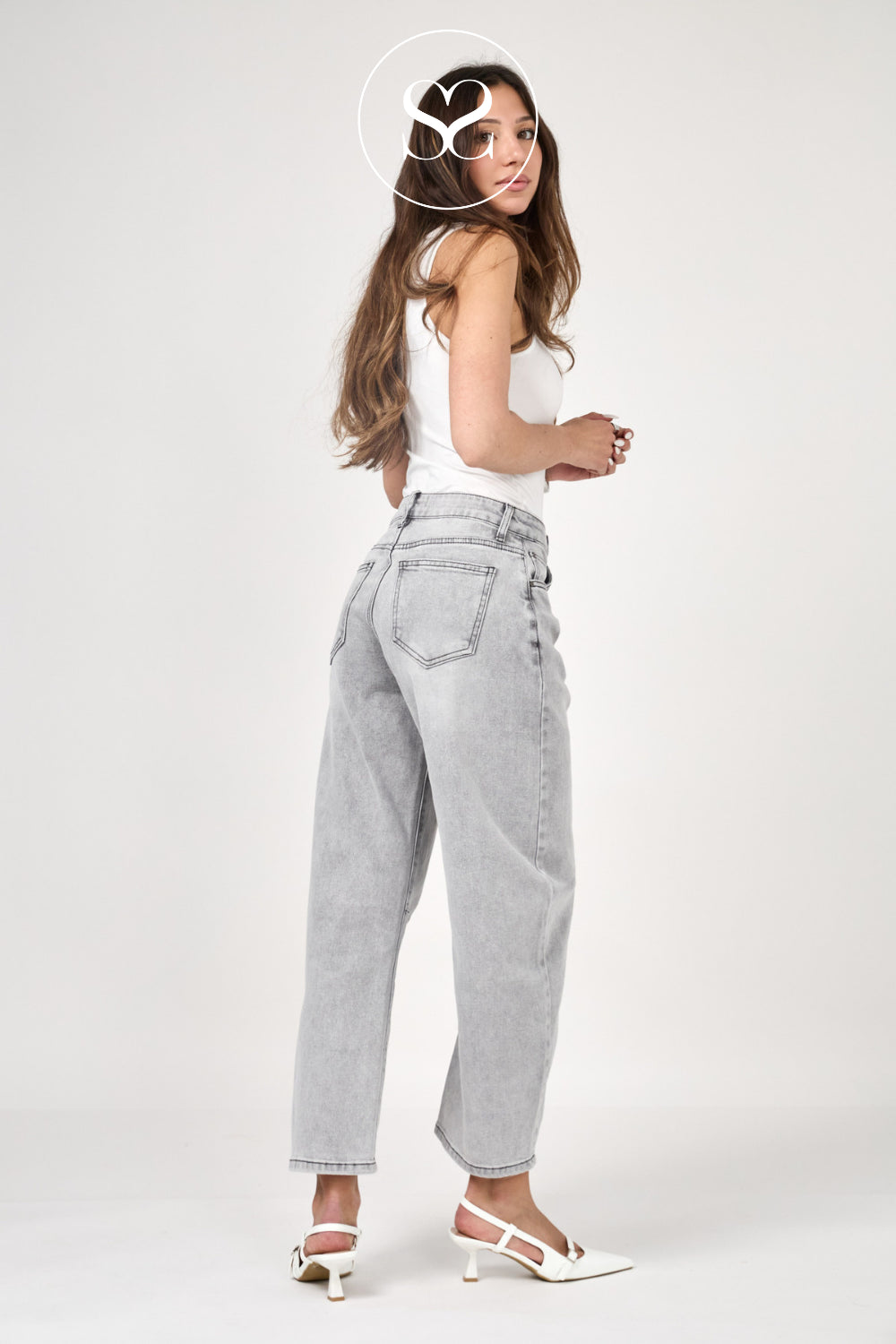 A woman modeling light-wash gray high-waisted straight-leg jeans, paired with a white tank top and white heeled sandals, photographed against a seamless light gray studio background.