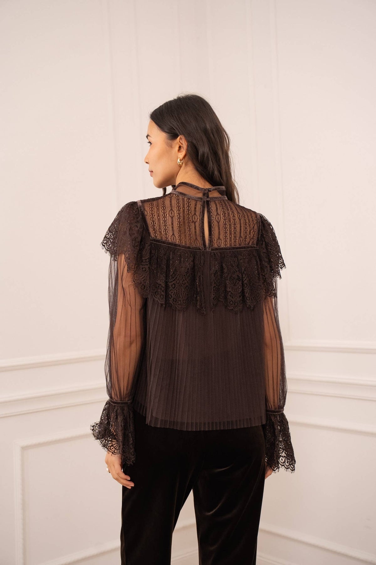 Woman wearing a dark brown blouse with lace details against a white wall.