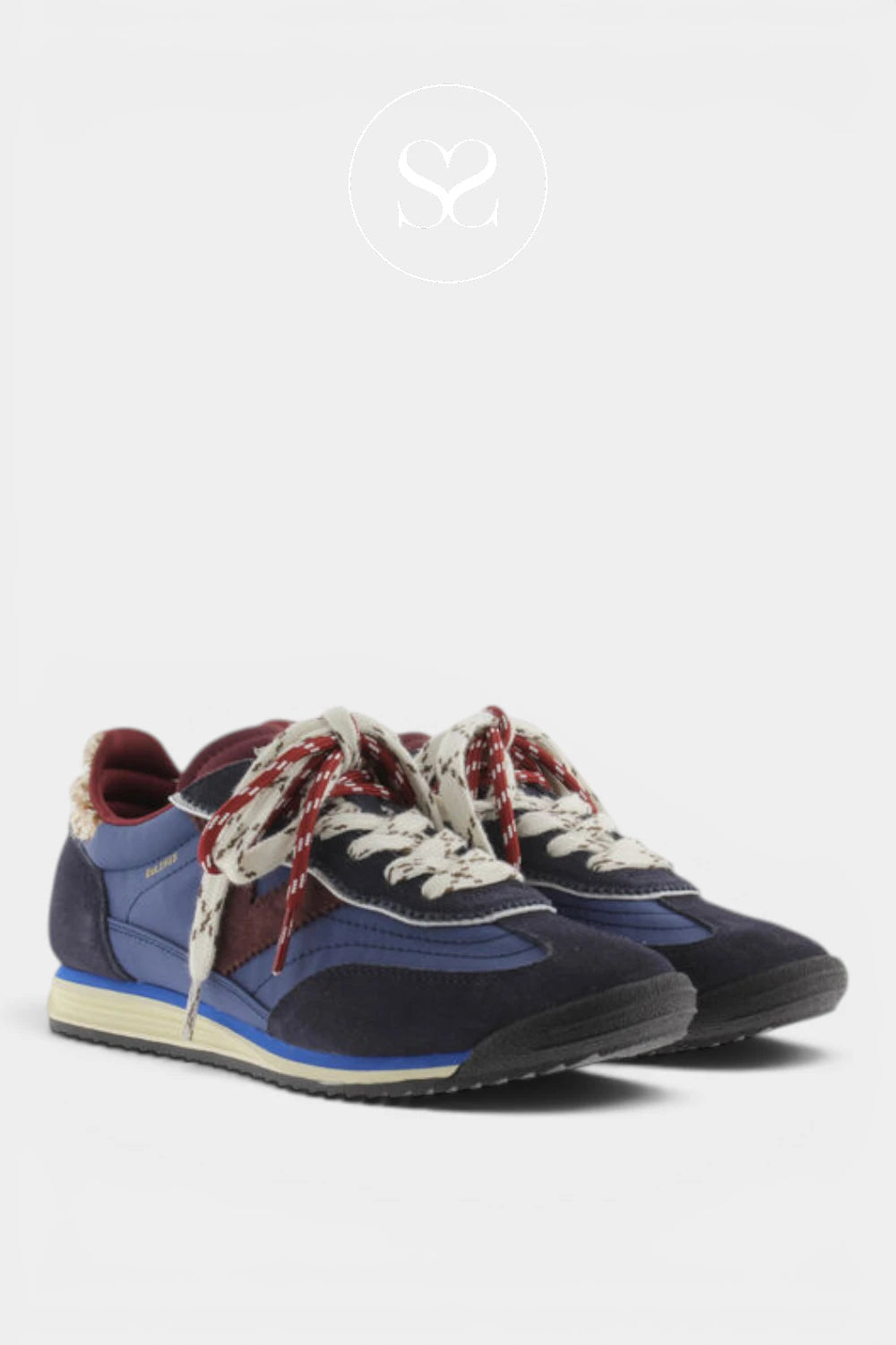 A pair of low-top sneakers in navy blue suede and canvas, featuring cream laces with red patterned details, a red inner lining, and a cream midsole, shown against a light gray background.
