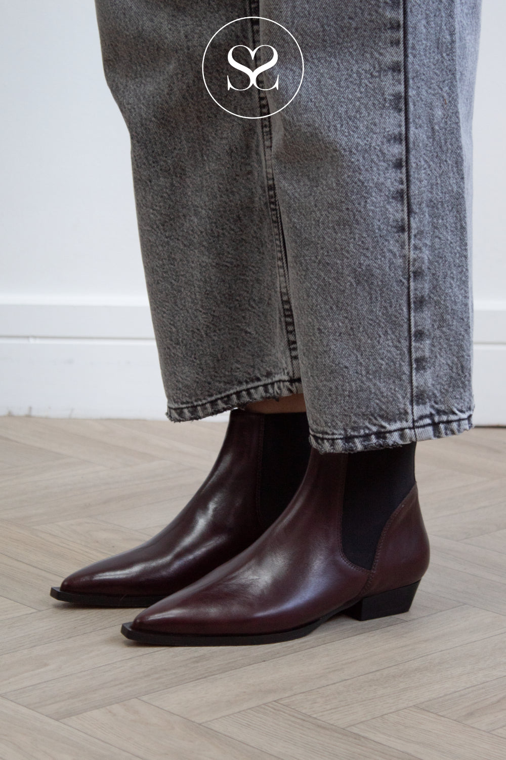 A woman wearing pointed-toe dark brown leather flat ankle boots with black elastic side panels, paired with gray jeans, standing on a light wood floor.