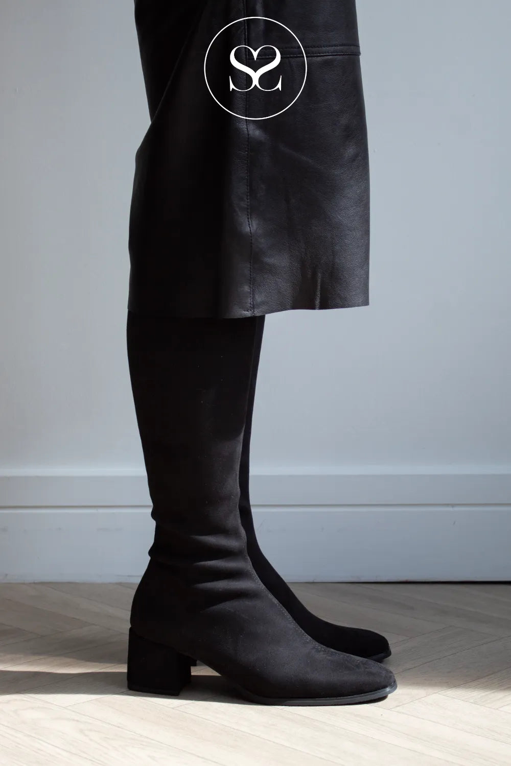 A person wearing black suede knee boots with a low block heel, paired with a black skirt, standing on a light wood floor against a gray wall. Miss elastic style pictured in Sheneil Shoes Ireland