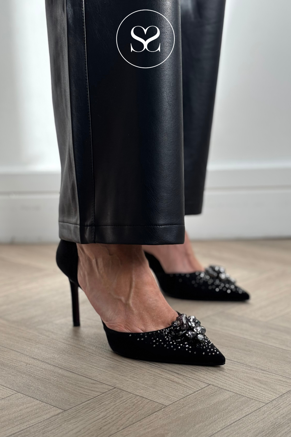 Black pointed-toe high heels with crystal embellishments, shown on a model wearing black faux-leather trousers on a light wood floor.