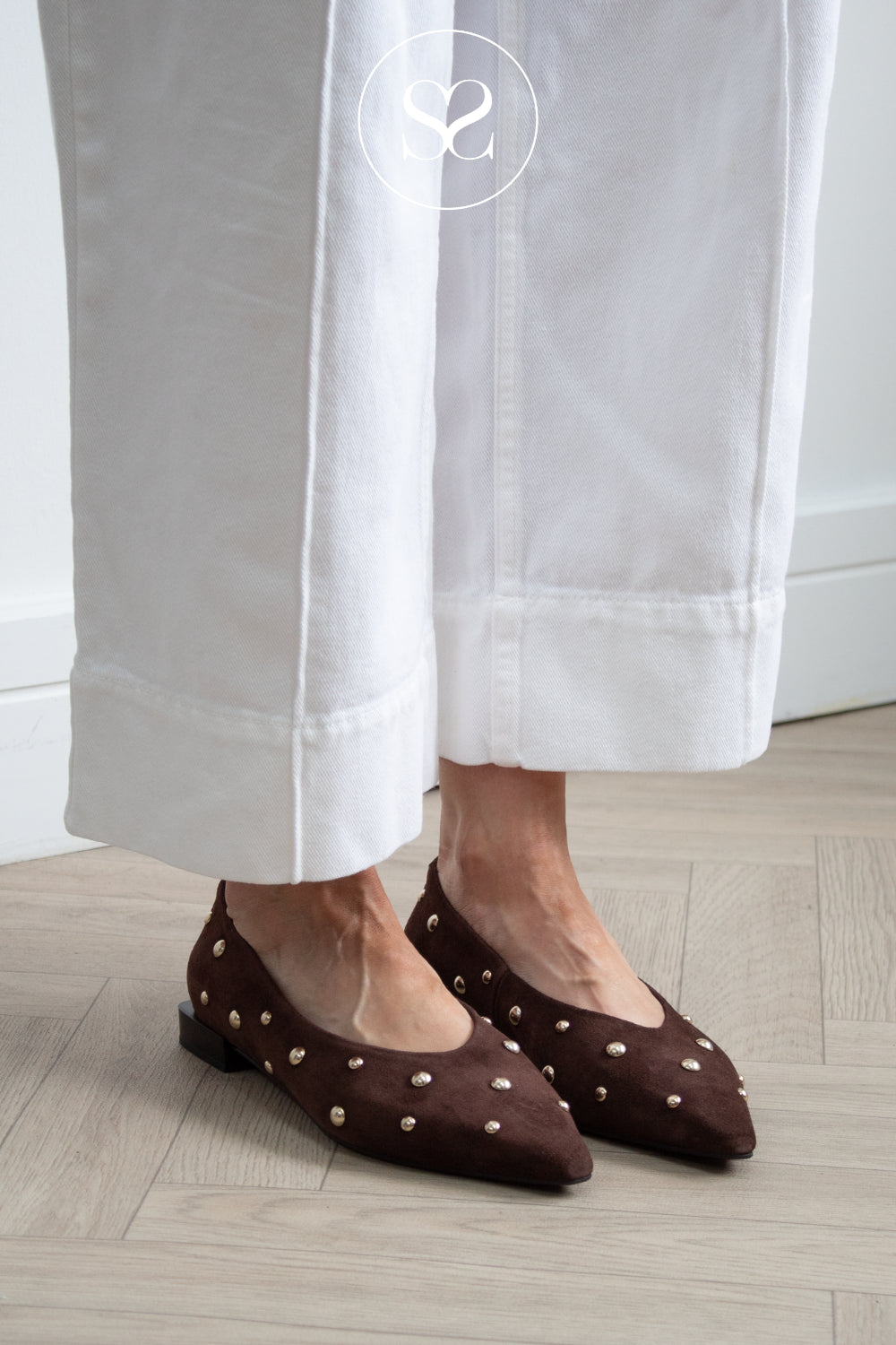 A person wearing dressy dark brown pointed-toe flats with gold-tone studs from Angel Alarcon, paired with white cropped pants, standing on a light wood herringbone floor.