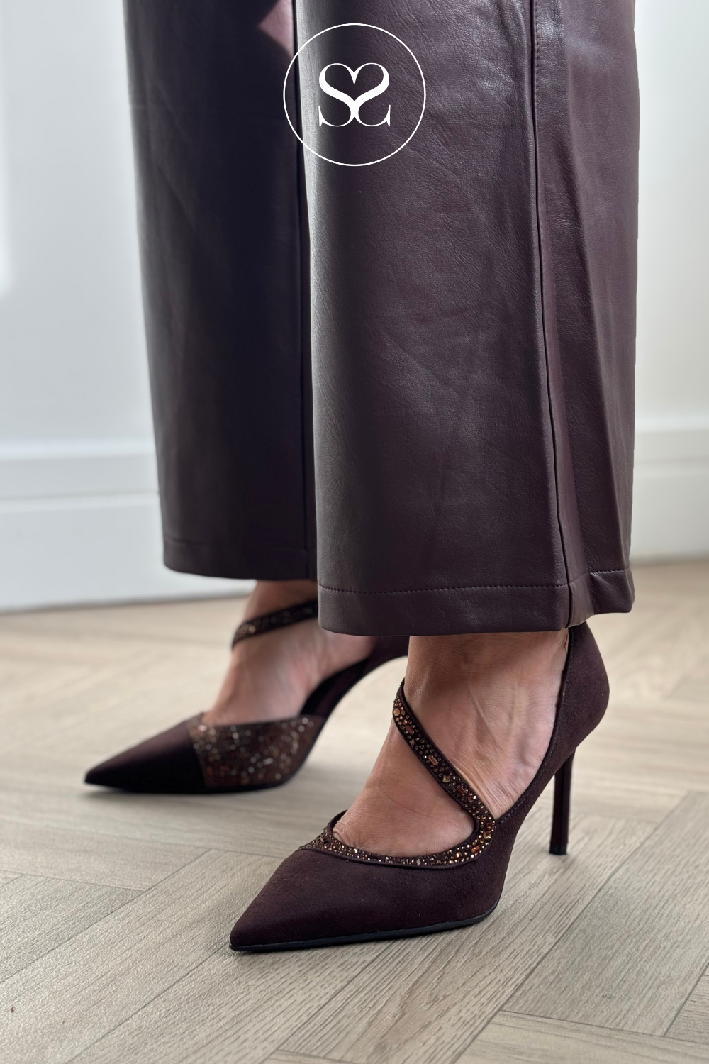 A person wearing dark brown pointed-toe high heels with glittery straps and matching dark brown wide-leg pants, standing on a light wood floor.