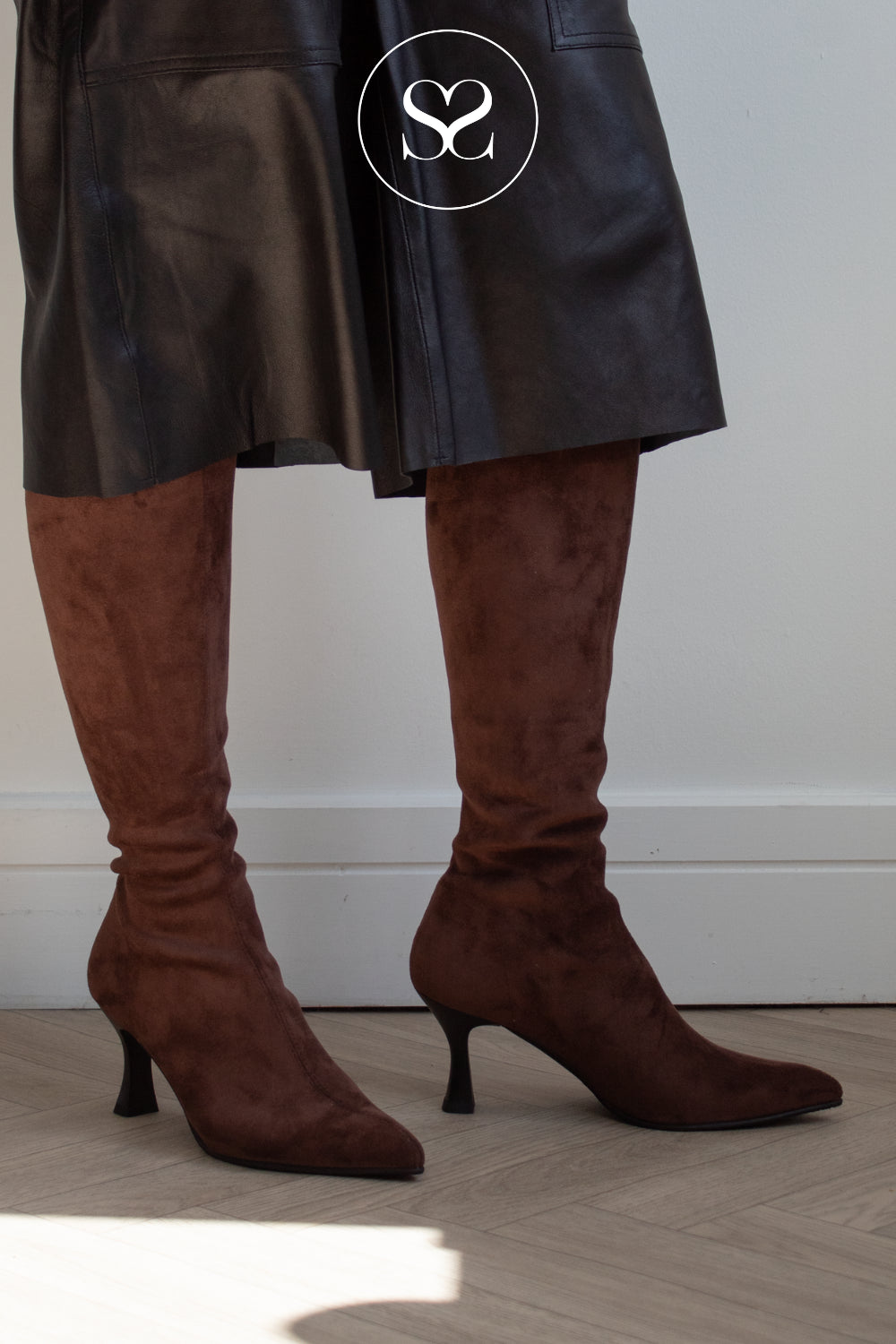 A person wearing dark brown knee high kitten heeled sock boots with a black skirt, standing on a light wood floor against a white wall.