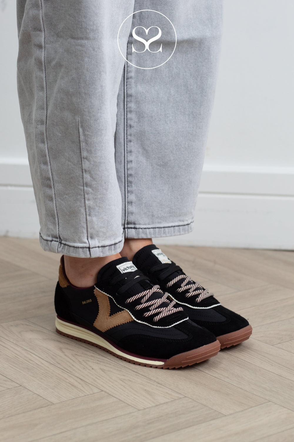 A person wearing black and tan suede sneakers with light gray jeans, standing on a light wood herringbone floor against a white wall.