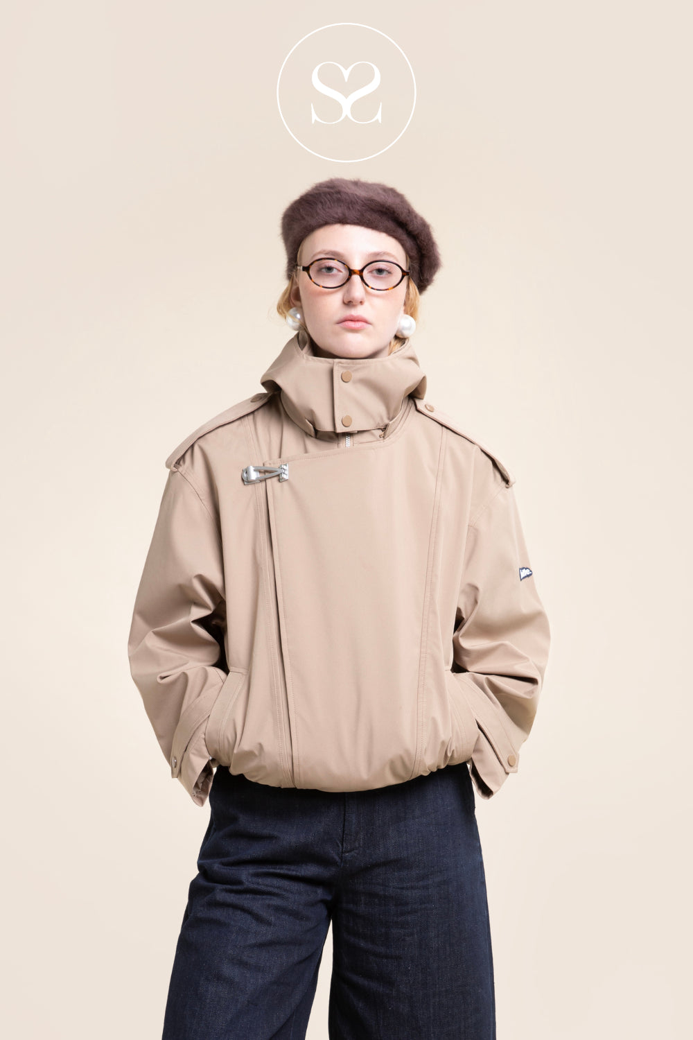 A model wearing a beige technical hooded jacket from Flotte with a high collar, paired with dark blue jeans and a brown beret, set against a solid beige background with a circular 'Sheneil Shoes' logo at the top.