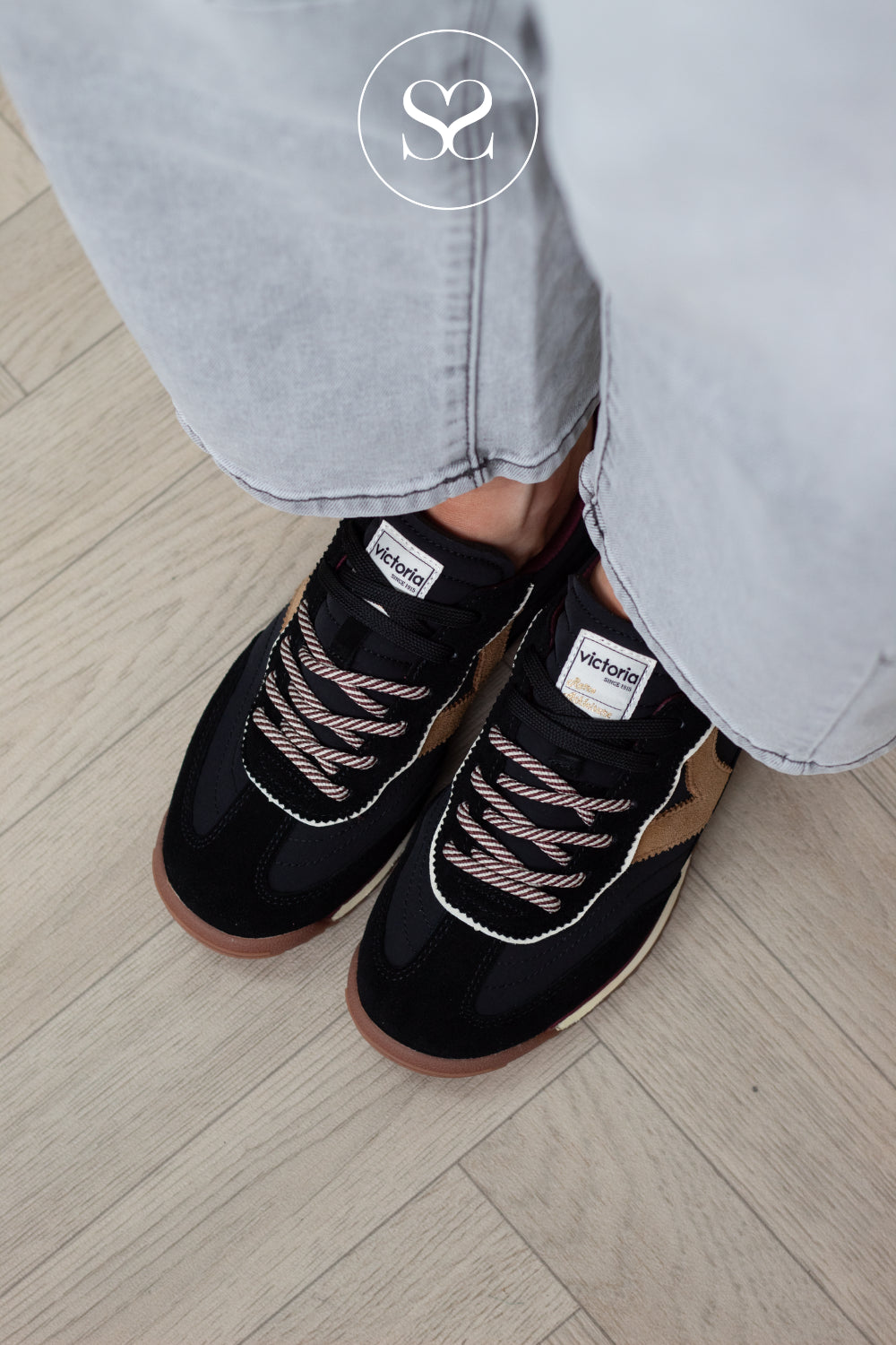 A close-up of a person wearing black Vichelin sneakers with brown and pinkish laces, paired with light gray jeans, standing on a light wood floor.