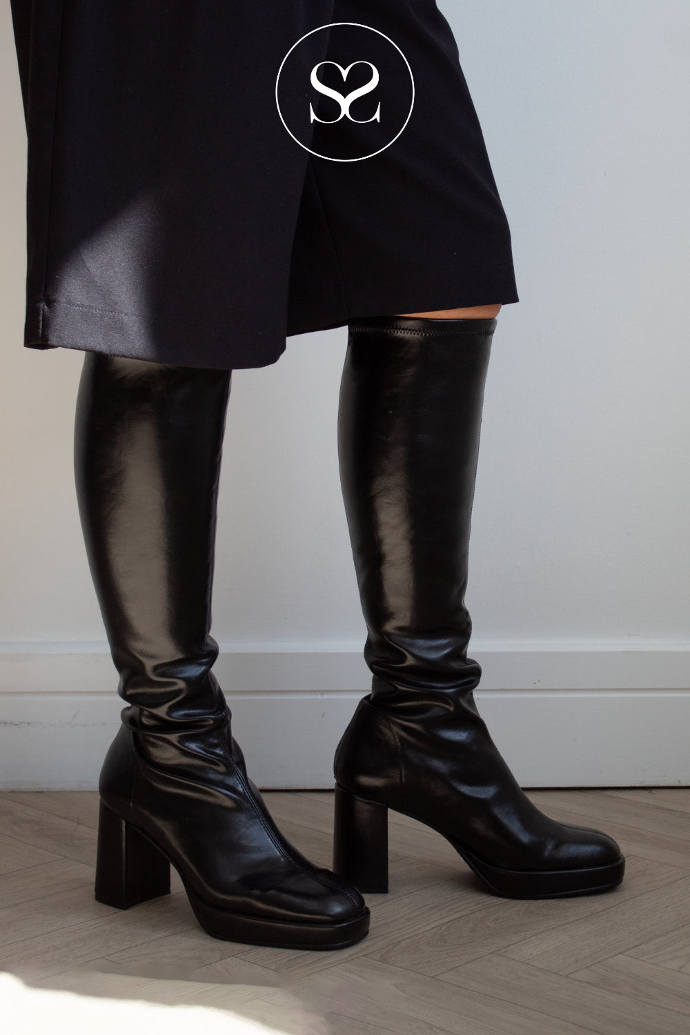 A woman wearing Miss Elastic shiny black knee-high heeled boots with a dark skirt, standing indoors against a light wall and wood floor. A circular 'Sheneil Shoes' logo is overlaid near the top.