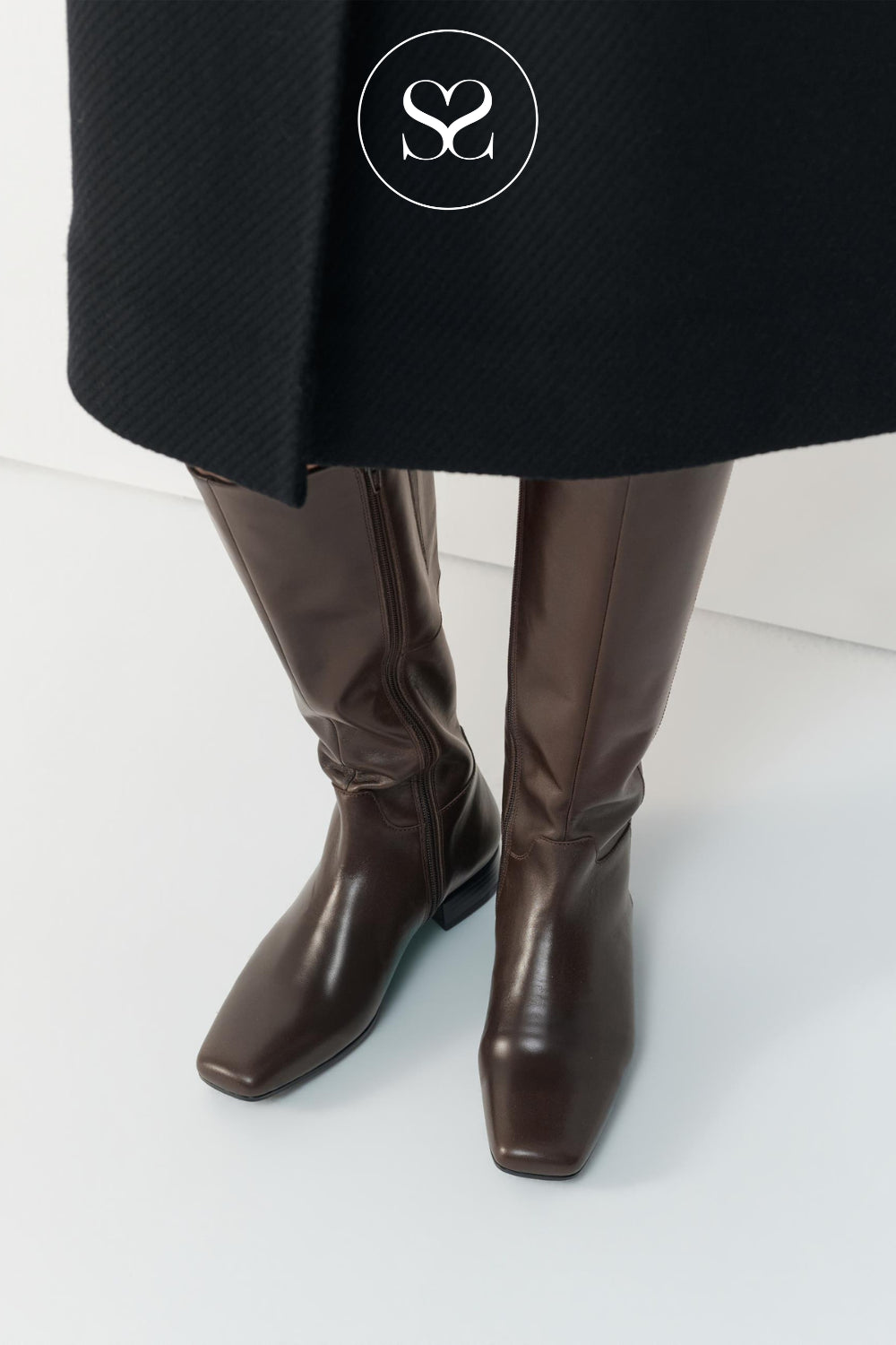 A close-up of a person's legs wearing dark brown leather knee-high boots with a black coat, featuring a white circular Sheneil Shoes logo overlay on the upper black coat.