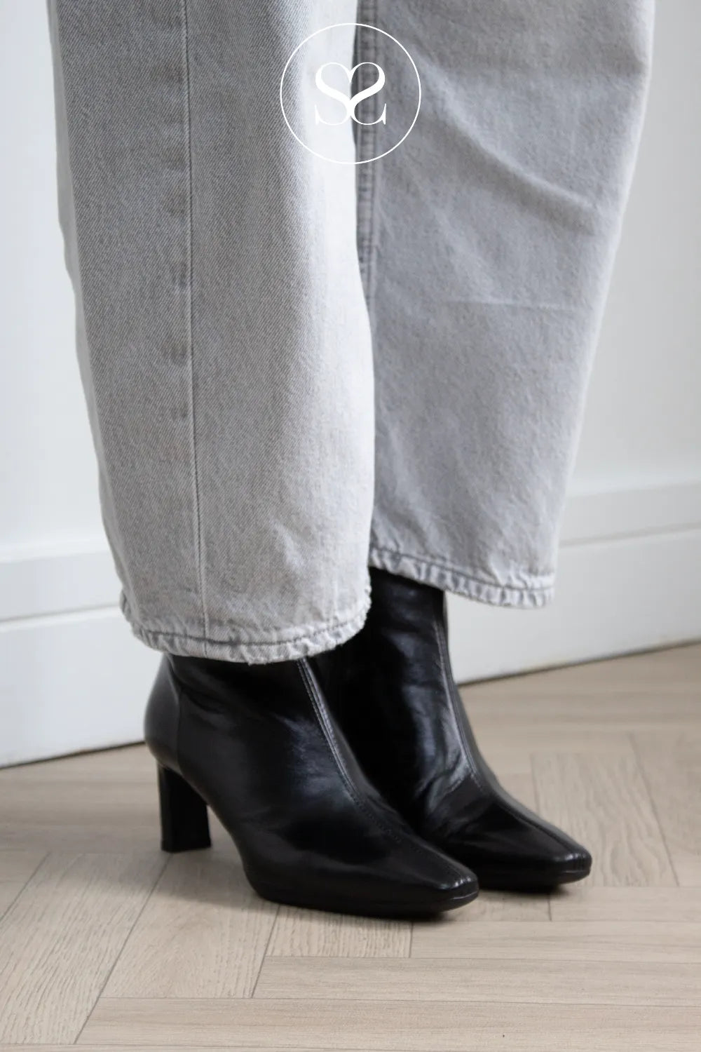 Wonders Black leather ankle boots with a block heel, worn with light grey jeans on a light wood floor. A white circular  Sheneil Shoes logo watermark is visible at the top.