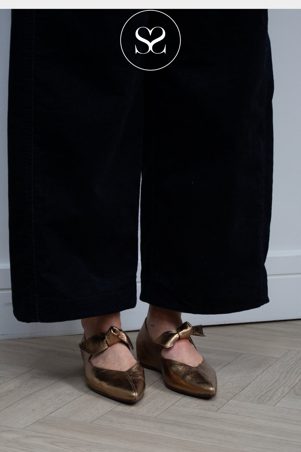 A woman wearing black wide-leg trousers and metallic bronze Mary Jane flats with bow details, standing on a light wood floor against a white wall. Shoes from wonders