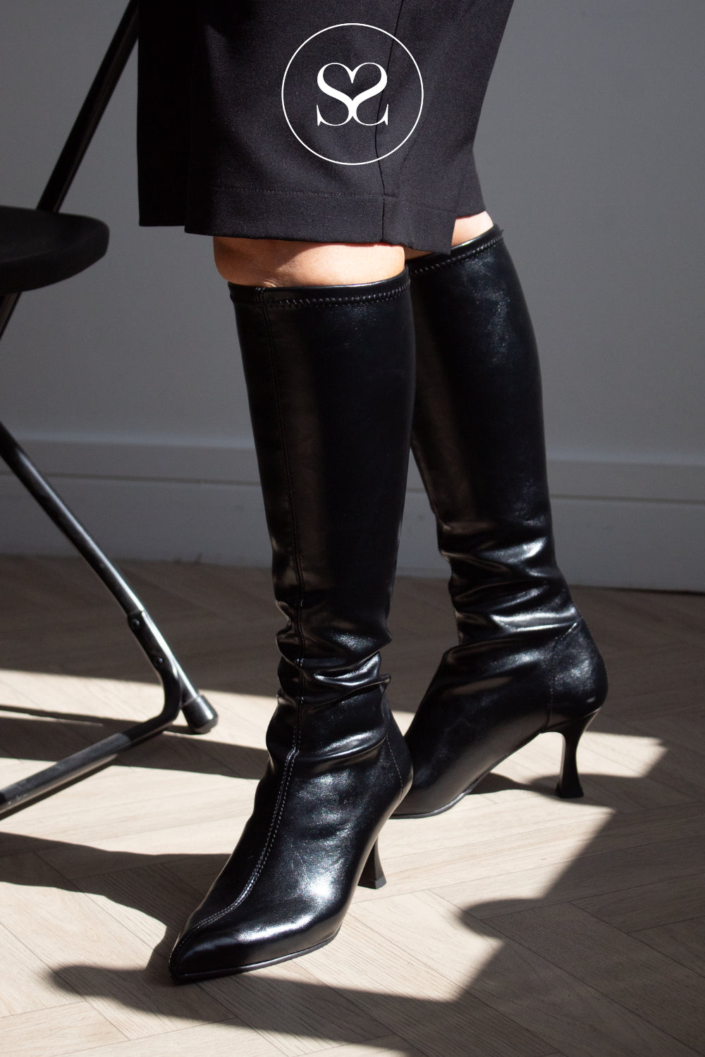 A person wearing shiny black knee-high heeled boots with pointed toes and kitten heel from Miss Elastic shown from the knees down in a real indoor setting with sunlight and shadows on a wood floor.