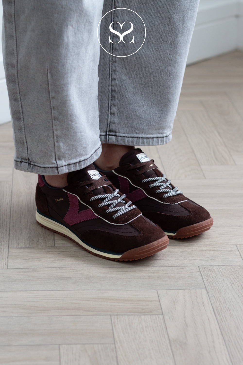 A person wearing dark brown suede sneakers with burgundy side panels, white rope laces, and light beige midsoles, paired with light gray jeans on a light wood herringbone floor.