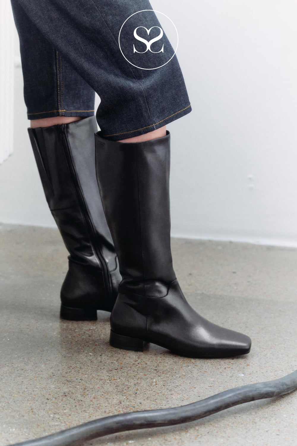 A person wearing Vagabond mona black leather knee-high boots with dark blue denim shorts, standing on a concrete floor against a white wall.
