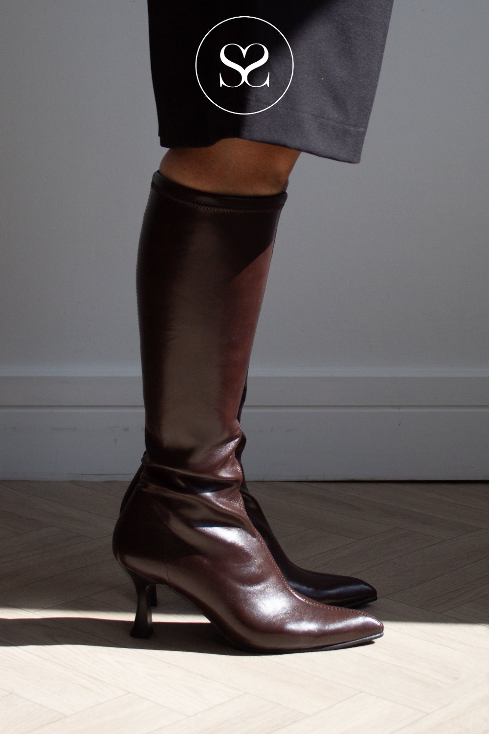 A pair of dark brown knee-high leather look sock boots with kitten heels, shown on a model wearing dark gray trousers. The photo is taken in a minimalist room with a light gray wall and herringbone wood floor, featuring a white circular 'SS' logo overlay near the top.