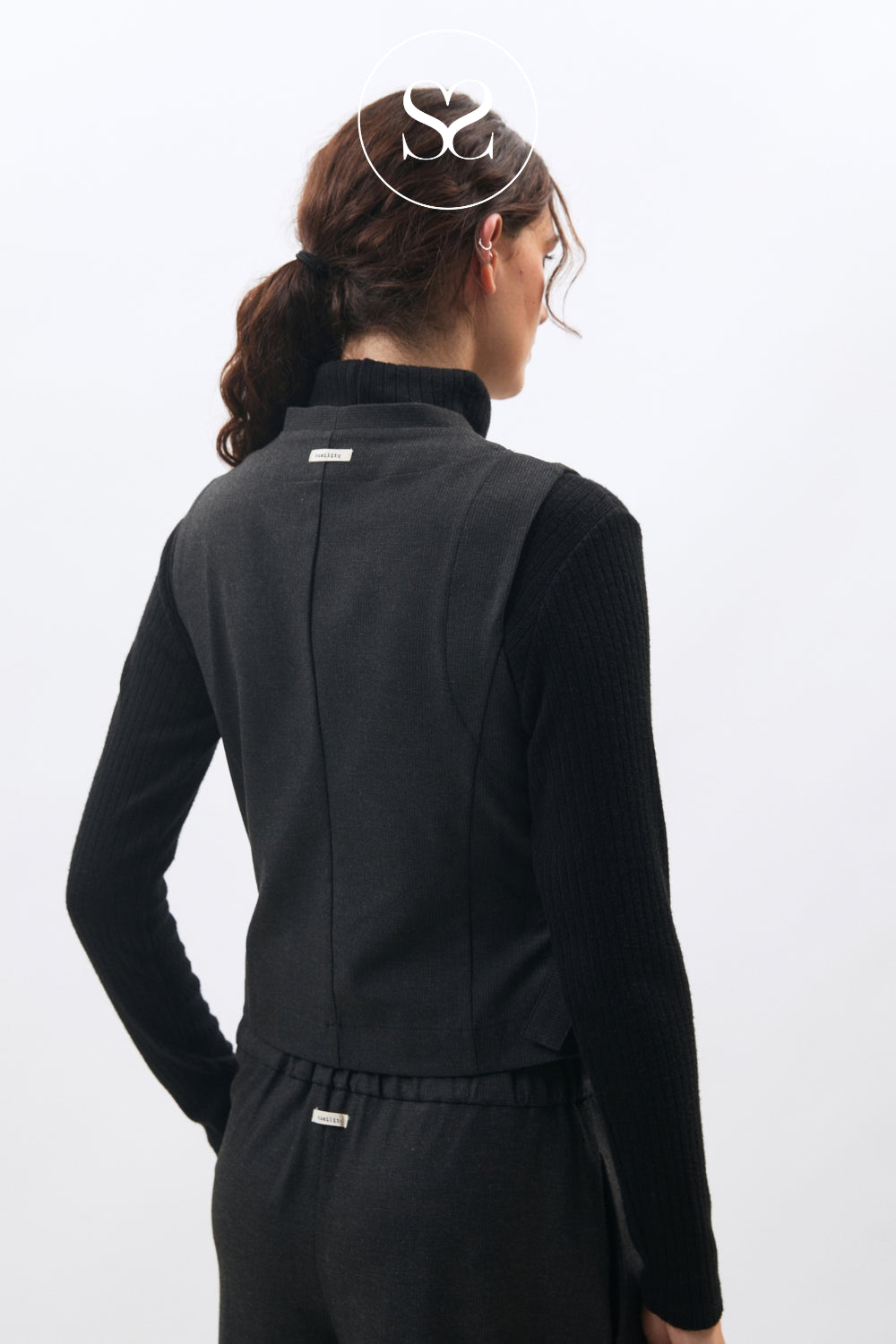 A woman shown from the back wearing a black ribbed long-sleeve top and matching black bottoms against a plain light gray studio background.
