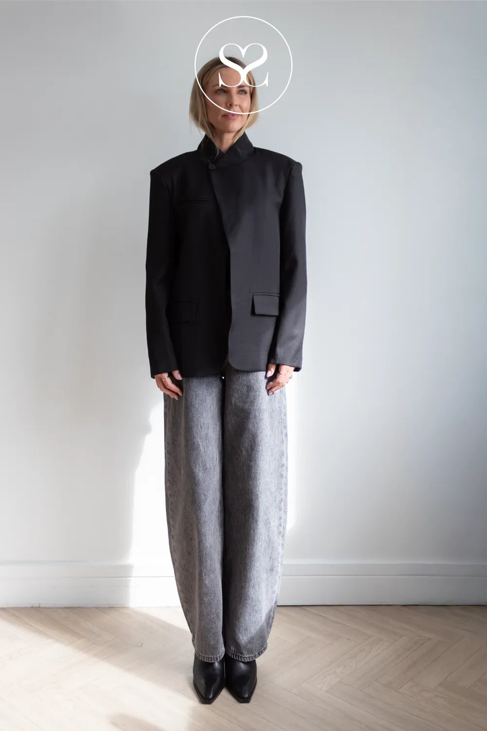A model stands in a minimalist room with a light gray wall and wooden floor, wearing a black asymmetrical jacket with a high collar, paired with loose gray trousers and black pointed shoes. A white circular logo overlay is positioned over the model's head.