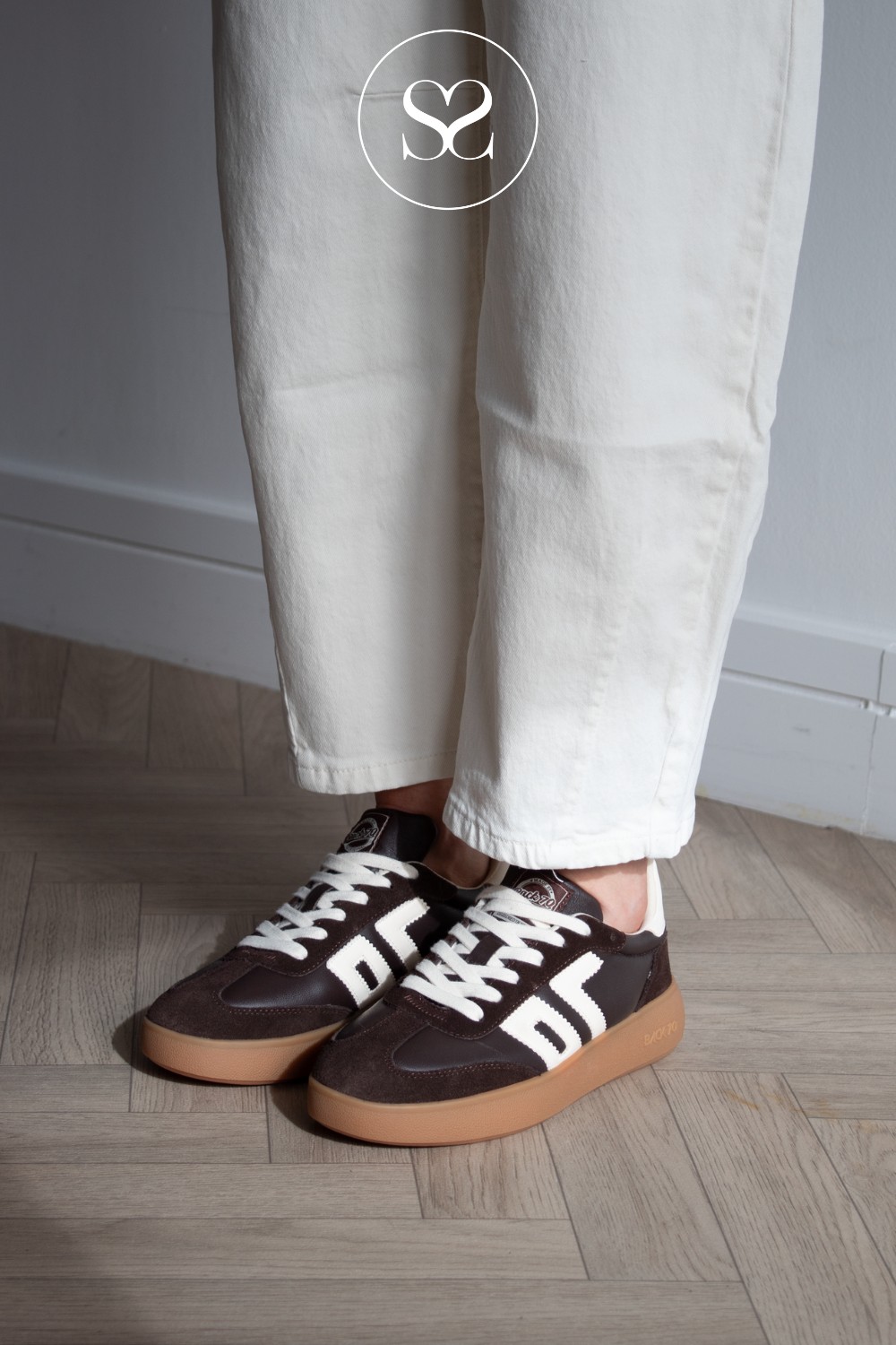 A person wearing brown leather low-top sneakers with white stripes and white laces, paired with off-white trousers, standing on a light wood floor.