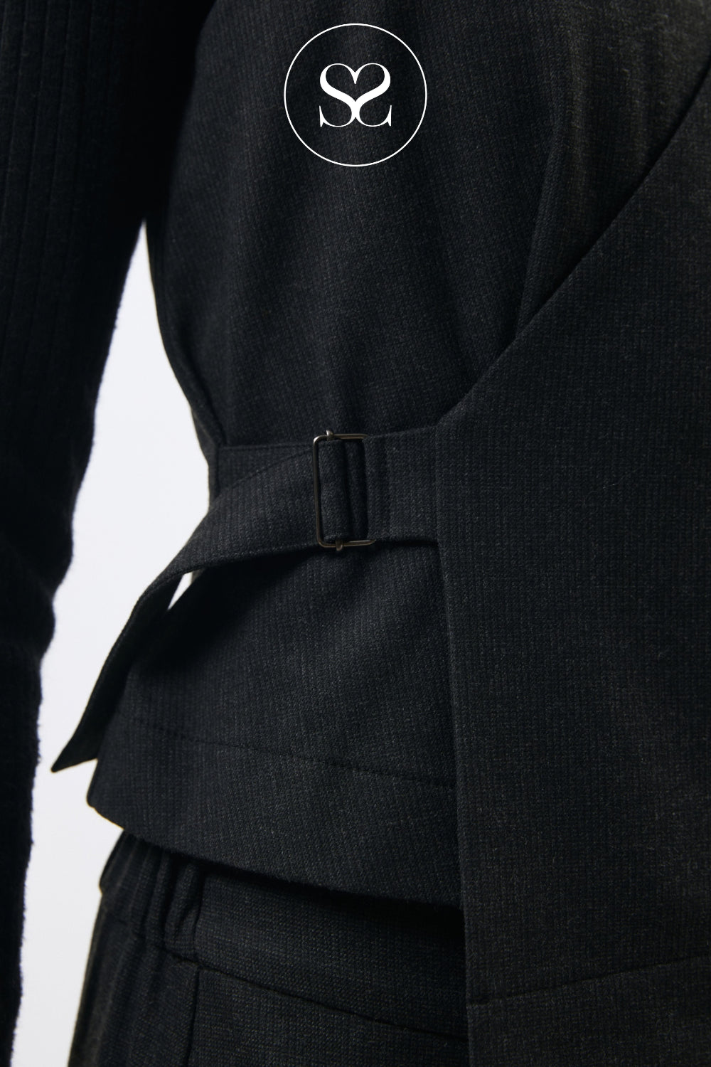 Close-up of a black ribbed knit garment with a wrap-style belt and buckle detail, featuring a white circular 'SS' logo near the top.
